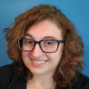 Jen Bauer Headshot, brunette with short curly hair, hazel eyes, blue glasses, smile